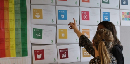 To elever, piger set fra ryggen, står og peger på en opslagstavle med FNs 17 verdensmål